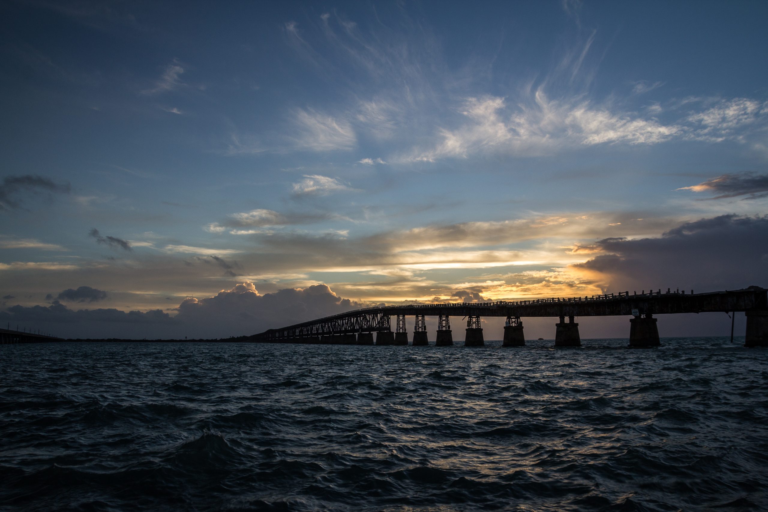 Silhouette of a bridge over water during sunset with cloudy sky.